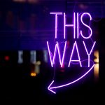 Purple neon light inside a restaurant stating: "This way" above an arrow pointing left.